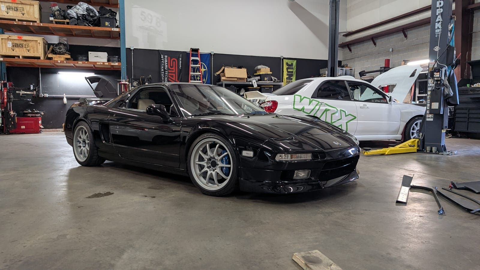 Black first-generation Acura NSX on the shop floor with a white Subaru WRX on the lift behind it